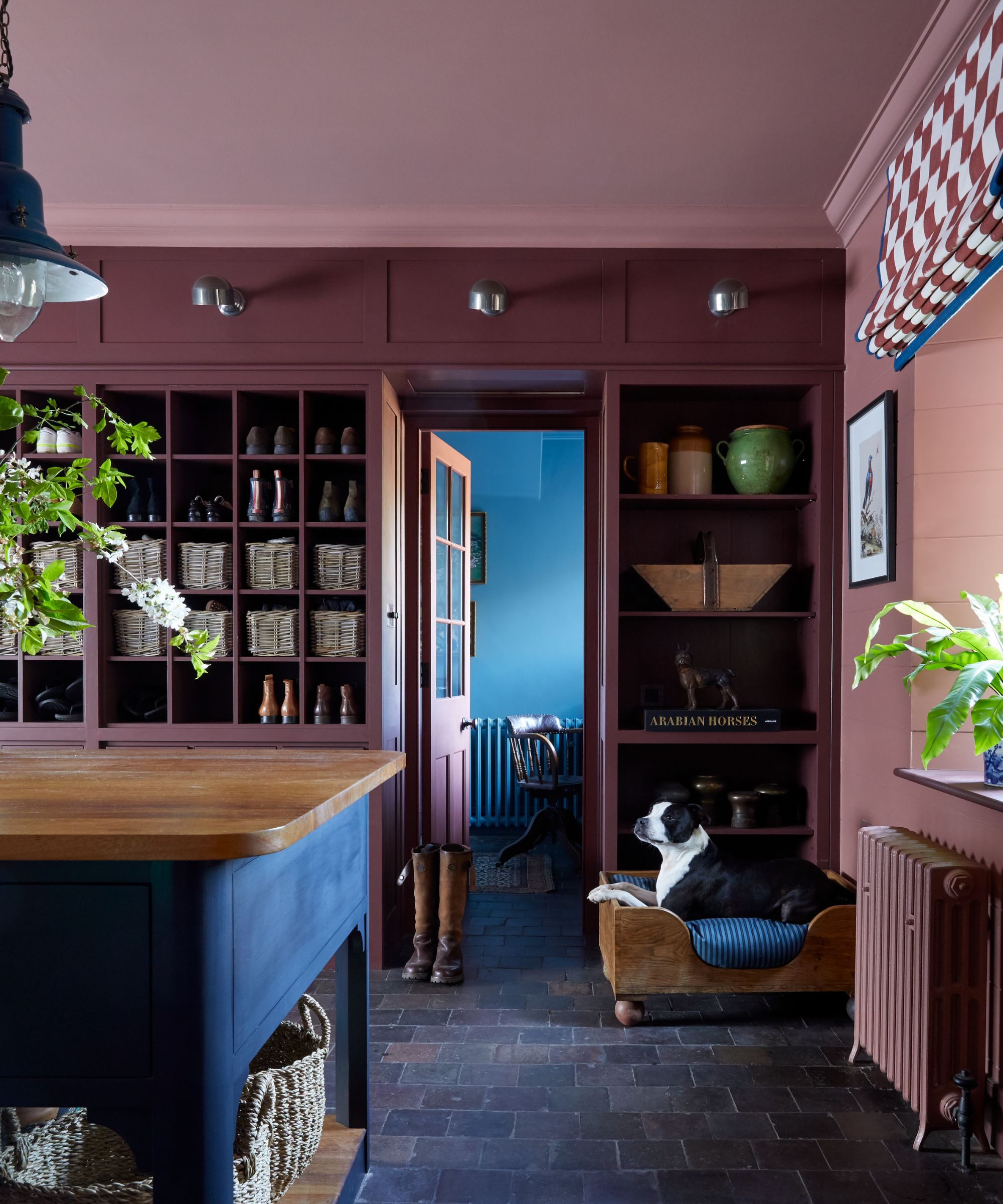 A rustic kitchen with a wall of shelving painted burgundy, a pink ceiling, a wooden kitchen island, and gray floors.