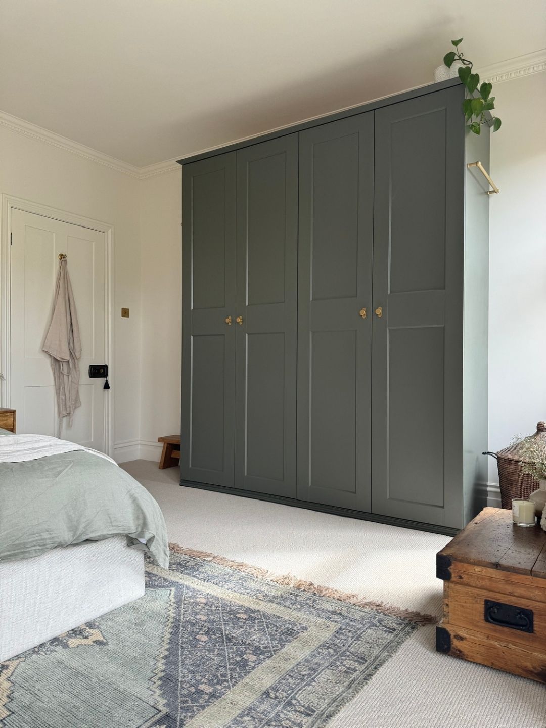 Image of a sage green wardrobe in a white room with off-white carpeting, a blue rug, and a bed with sage green bedding.