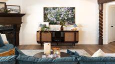 Living room with vintage media unit below a framed floral photograph. A blue sofa is pictured in the foreground, alongside wooden beams, a round coffee table, a gray rug, a fireplace, and wooden floors.