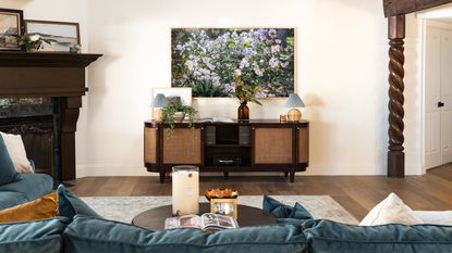 Living room with vintage media unit below a framed floral photograph. A blue sofa is pictured in the foreground, alongside wooden beams, a round coffee table, a gray rug, a fireplace, and wooden floors.