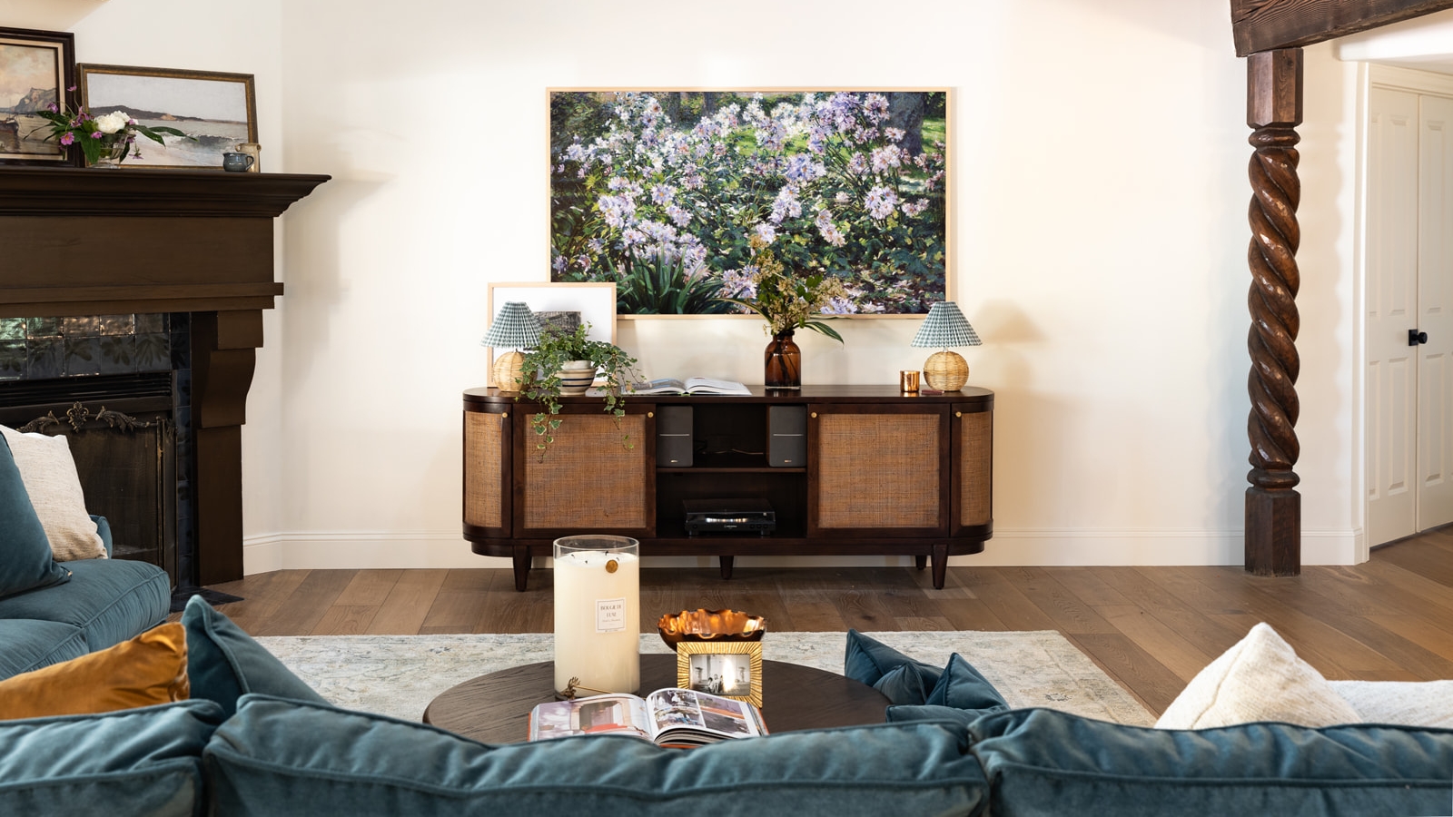 Living room with vintage media unit below a framed floral photograph. A blue sofa is pictured in the foreground, alongside wooden beams, a round coffee table, a gray rug, a fireplace, and wooden floors.
