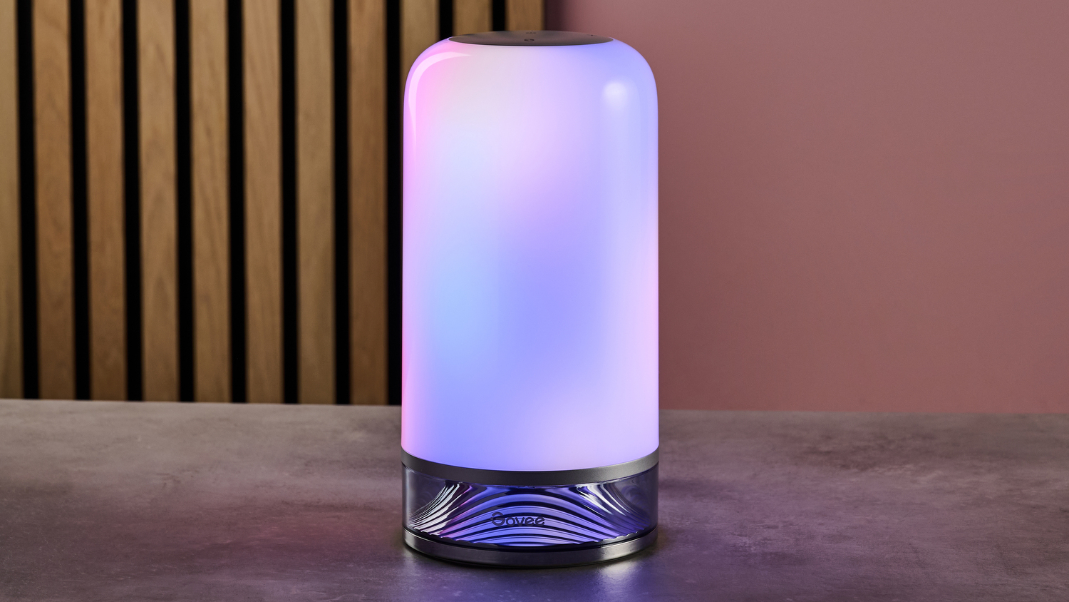 The Govee Table Lamp 2 sits on a stone-effect table in a dimly lit pink room. It is displaying a pretty mixture of blues, pinks, and purples.