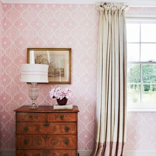 A pink room with a window and a curtain next to a wooden dresser