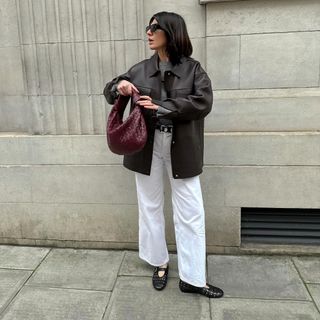 woman wearing leather jacket, white jeans, black studded ballet loafers, and carrying a burgundy bag