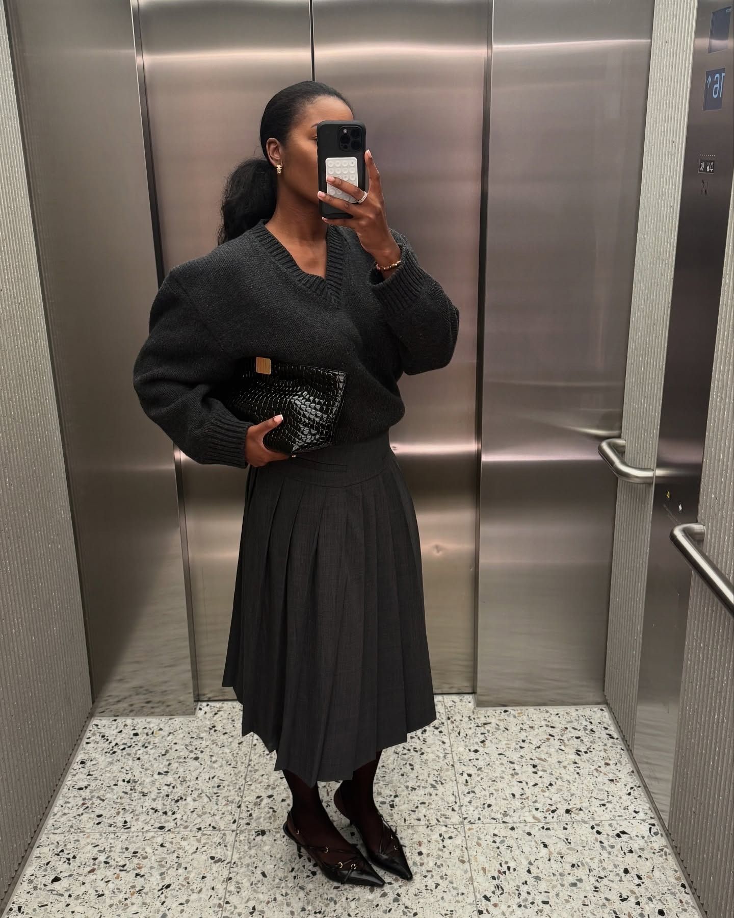 Marilyn wears a gray sweater, black croc bag, gray skirt, and black slingbacks.