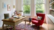 A bright home office with large windows, a wooden desk and a plush red armchair with patterned cushion. On the desk are piles of books and a table lamp. Behind it is a wheelie grey chair. On the wall are three framed artworks. On the opposite wall is a large bookshelf and white cabinet. The floor is wooden, with a patterned rug in the center.