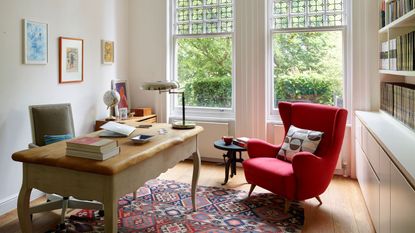 A bright home office with large windows, a wooden desk and a plush red armchair with patterned cushion. On the desk are piles of books and a table lamp. Behind it is a wheelie grey chair. On the wall are three framed artworks. On the opposite wall is a large bookshelf and white cabinet. The floor is wooden, with a patterned rug in the center.