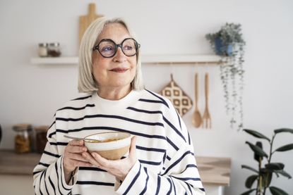 Elderly woman enjoying nutritious lunch, smiling warmly while seated in sunlit contemporary kitchen with clean design