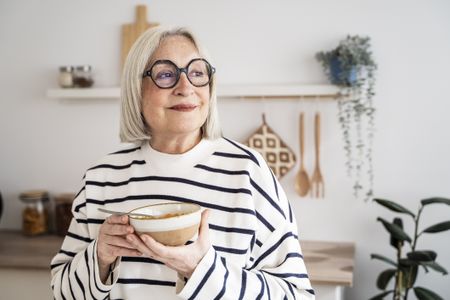 Elderly woman enjoying nutritious lunch, smiling warmly while seated in sunlit contemporary kitchen with clean design