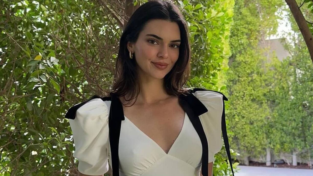 Kendall Jenner stands in front of landscaping wearing a white dress with black bows on the shoulders