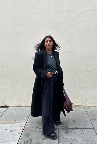 a photo showing what shoes to wear with wide-leg pants with a woman standing against a white backdrop while wearing a black long coat over a gray t-shirt with a studded black belt, black wide leg jeans, black chunky Chelsea boots, and a red Loewe bag