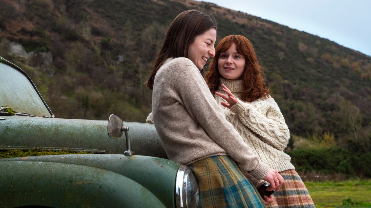 Hazel Doupe as Marian Price and Lola Petticrew as Dolours Price, leaning against the front of an old green car covered in dirt, in &#039;Say Nothing.&#039;