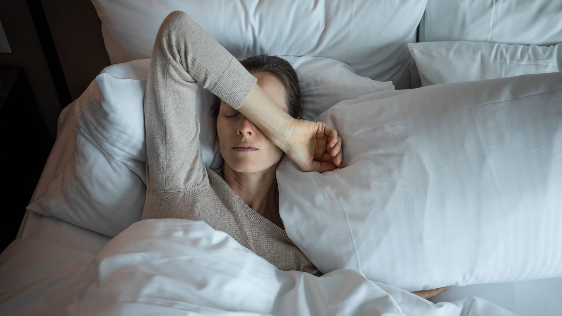 A woman lies in bed tired with her eyes closed and her arm over her face.