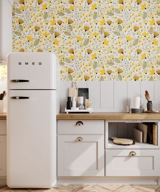A kitchen with yellow floral wallpaper, a white fridge, white cabinets, and a wooden countertop with white and black bottles, cups, and trays on it