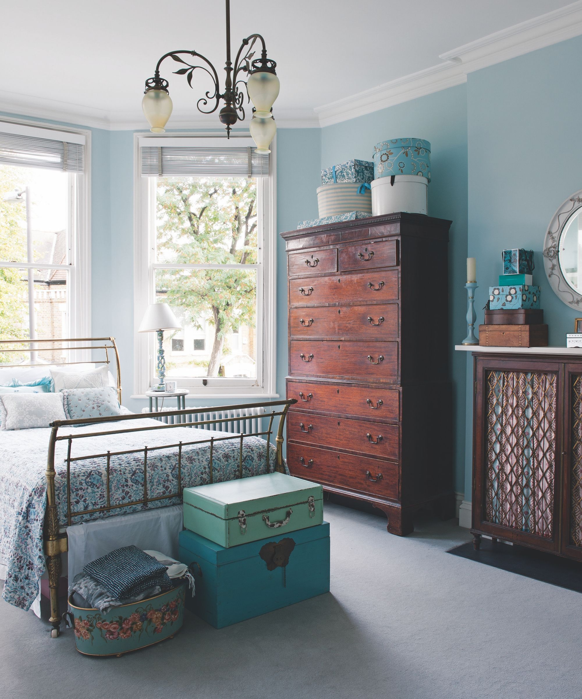 Blue guest bedroom with bay windows, a brass-framed bed, stacked colorful trunks, and a large dark wood chest of drawers.