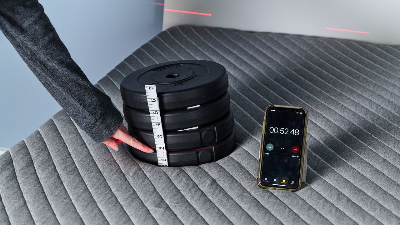The image shows a close up of a 10kg weight stack balanced on the shoulder area of the Siena Mattress in our sleep studio. A hand points to a tape measure on the side of the weight stack, noting that a laser is hitting the measure at roughly 1.5 inches. A phone with a timer is balanced to the right of the weight stack, showing a time of 52 seconds &mdash; that's how long the weight has been balanced for. The amount the weight has sank into the mattress indicates how much contouring you can expect from the Siena Mattress.