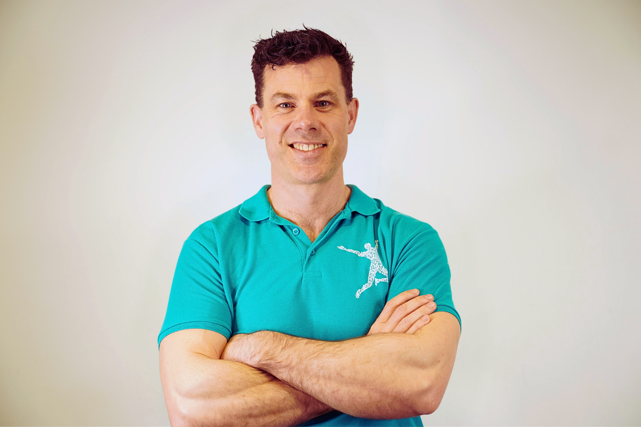 Head and shoulders of smiling man with arms crossed, wearing a teal polo shirt, standing against a blank studio backdrop