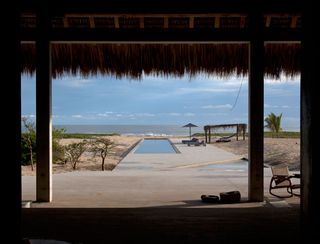 looking out from a thatched building towards a long swimming pool and the ocean