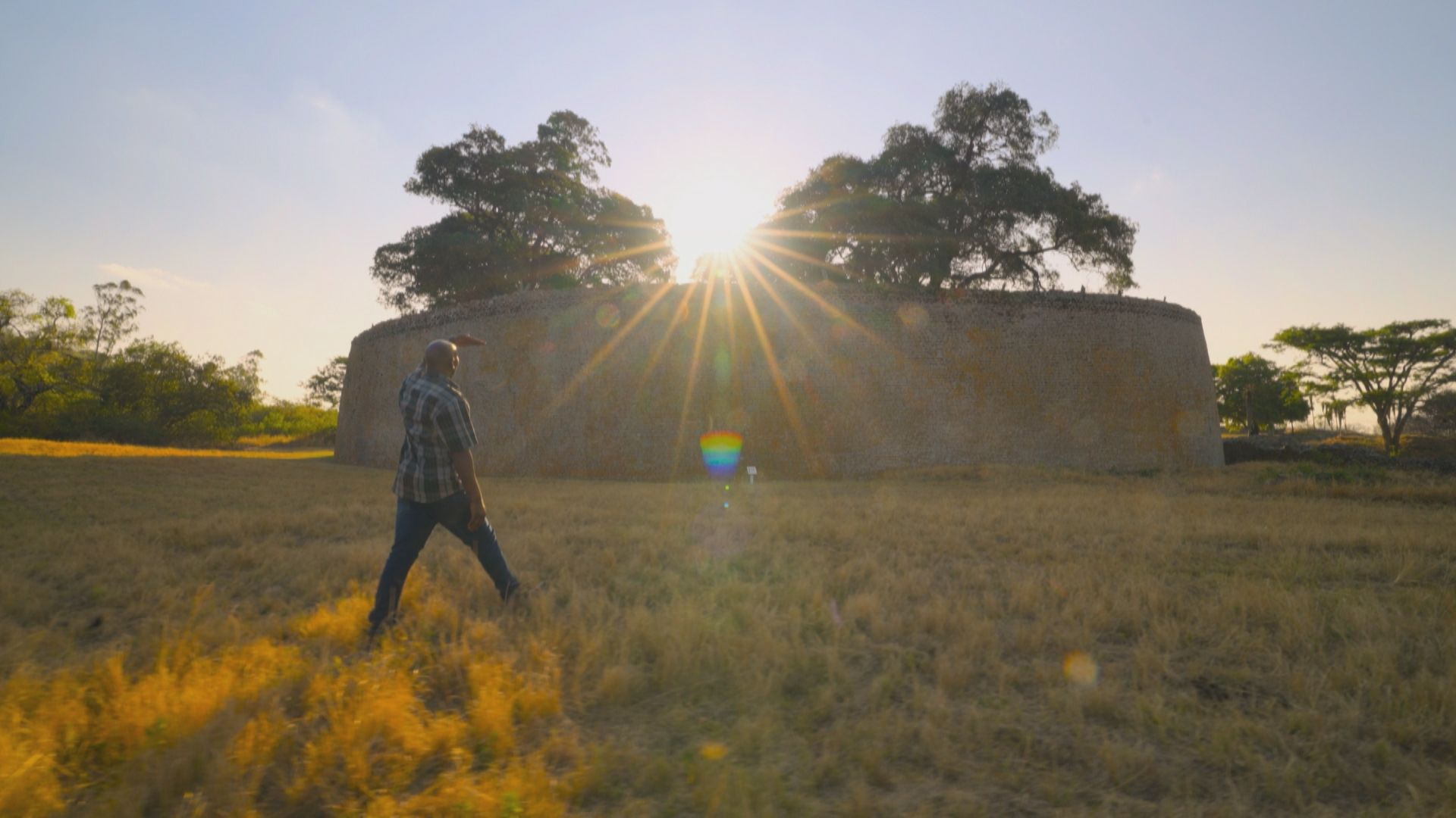a man walks across a grassy field with a circular stone enclosure rising in the background