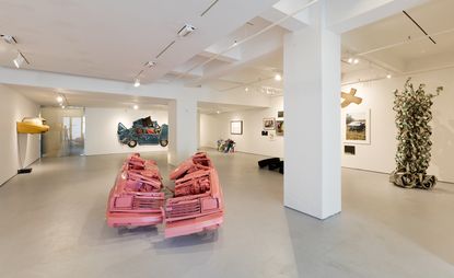 An art gallery featuring mangled parts of different cars on the floors and the grey floors and white walls of the room. 