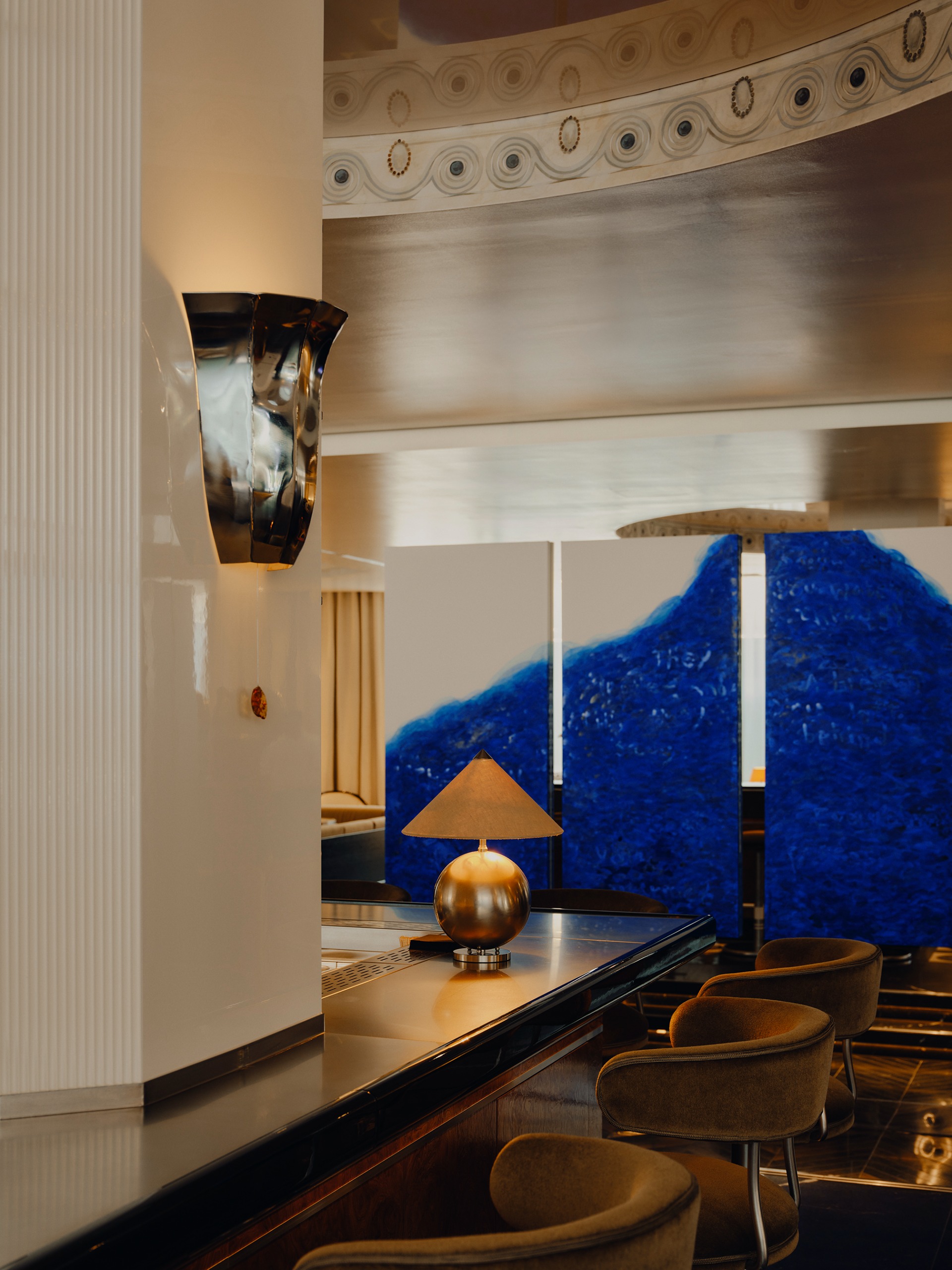 A bar with a stainless steel wall light and table lamp with a cone linen shade, with curved ochre and metal bar stools around it and a three-segmented artwork behind featuring bold blue against white