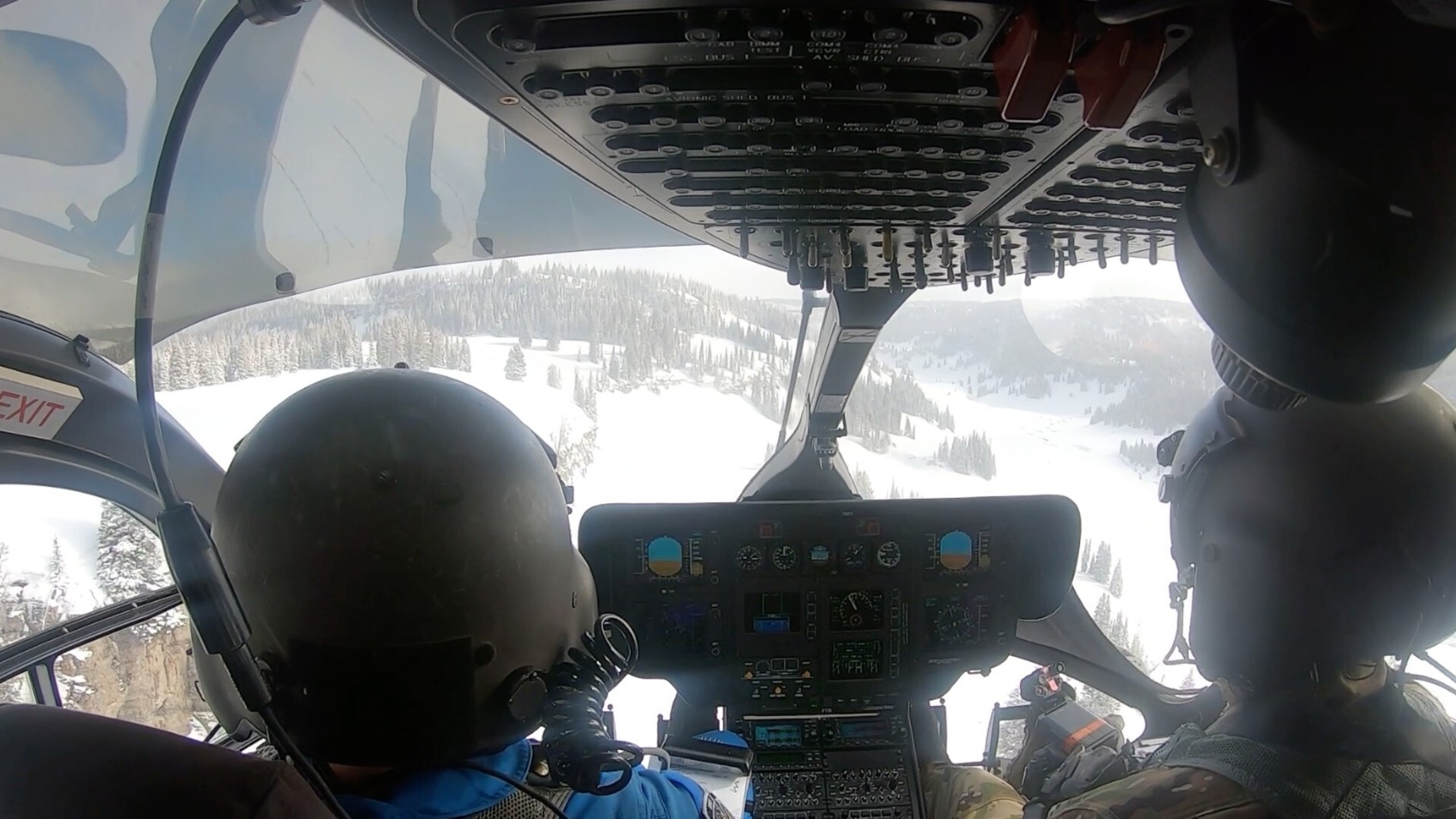 Two pilots sit in the cockpit of a plane wearing black helmets and flying a plane over some snowy mountains