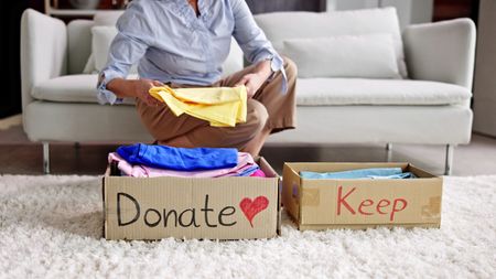A woman folds a shirt while sitting on the sofa behind boxes marked "donate" and "keep."