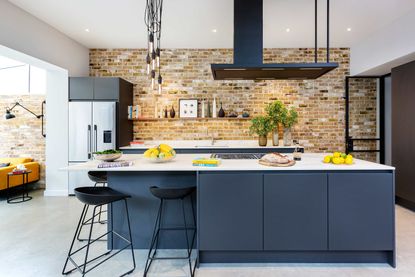 blue kitchen with exposed brick wall, white countertops, gray tiled floor, view into living space, bar stool, breakfast bar, pendant lights, modern cooker hood 