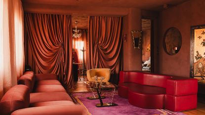 theatrical living room with orange plastered walls, pink carpet, red leather sofa, red velvet sofa, dramatic drapery over the doorway, and orange seat, and steel and glass coffee table