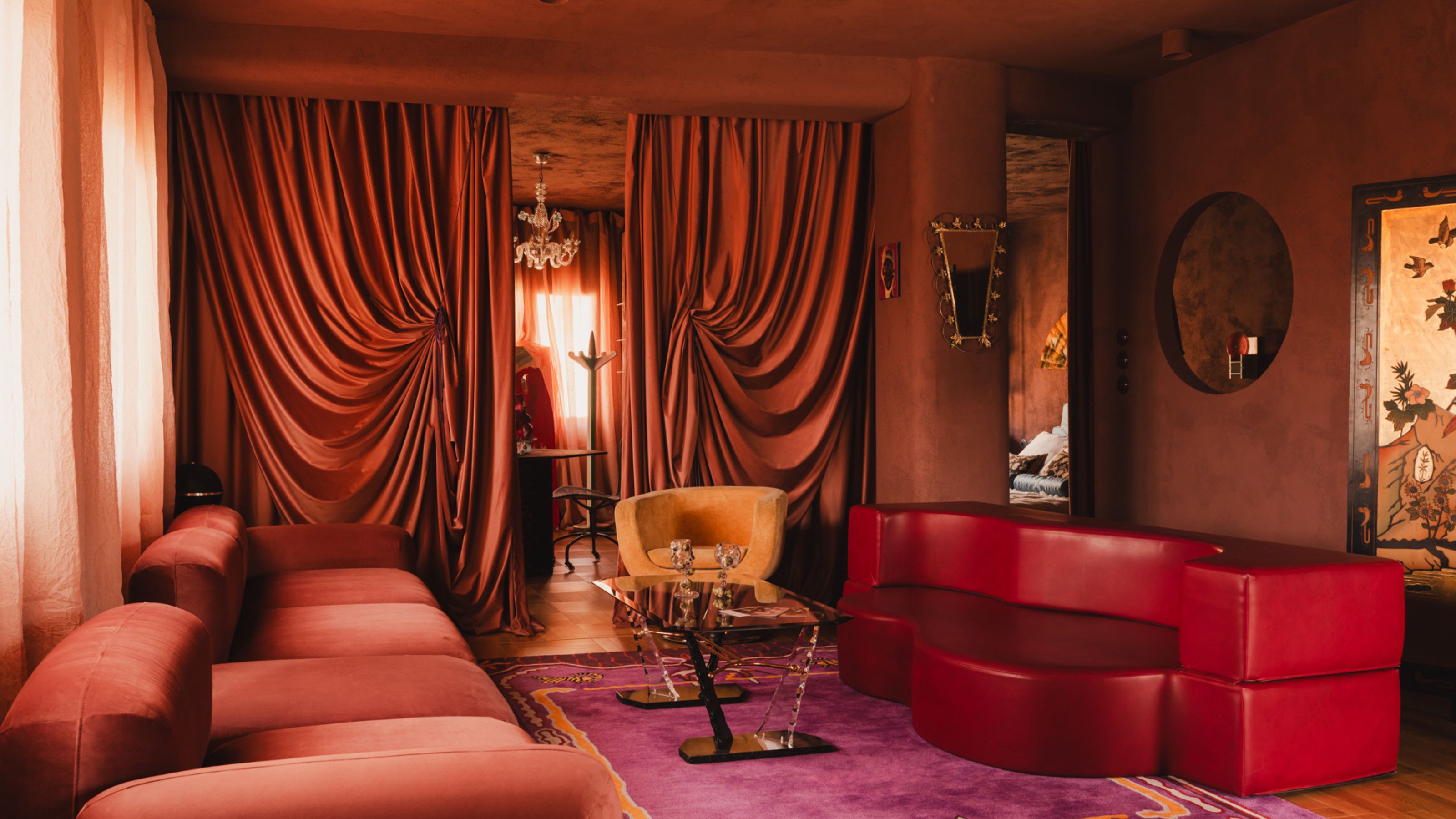 theatrical living room with orange plastered walls, pink carpet, red leather sofa, red velvet sofa, dramatic drapery over the doorway, and orange seat, and steel and glass coffee table