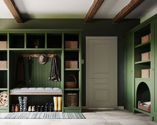 Large boot room with bespoke green wall storage and shelving unit, painted green walls, white painted ceiling with wooden beams, stone tiled flooring with rug on floor, white door