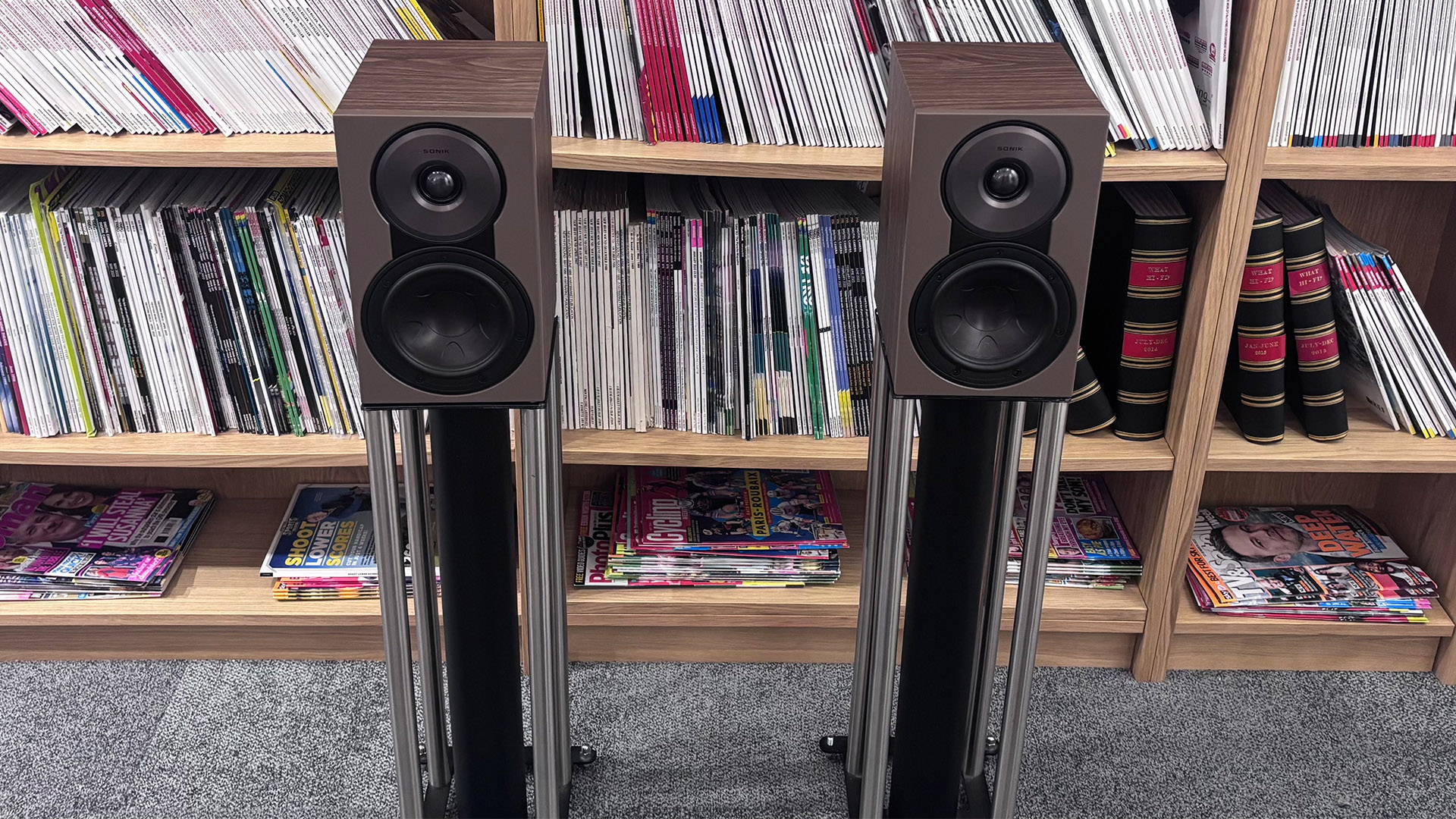 Dali Sonik 1 standmount speakers on stands on grey carpet in front of bookcase