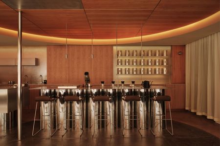 A theatrical cafe interior features backlit ceilings, white curtains, a chrome-clad, reflective bar counter, and chocolate leather stools with silver legs.