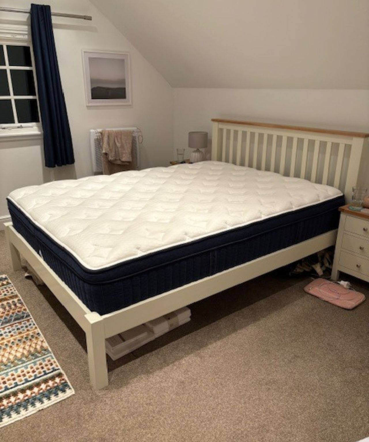 The Brooklyn Bedding Mattress shown on a white bed in a white attic bedroom with white matching bedside tables, beige carpet, and a colorful rug.