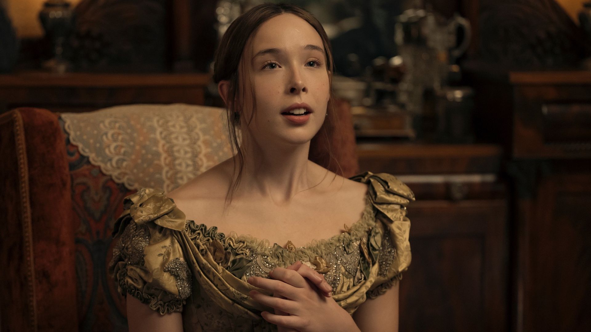 Lizzie Borden clutching her hands to her chest in awe during Episode 3 of Monster: The Lizzie Borden Story. She's wearing a green dress and seated on a red chair.