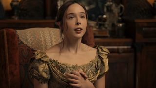 Lizzie Borden clutching her hands to her chest in awe during Episode 3 of Monster: The Lizzie Borden Story. She's wearing a green dress and seated on a red chair. 