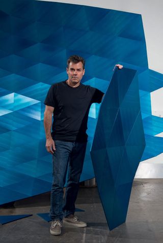View of Alex Rasmussen standing in front of 'The Wave' installation that he is working on. He is wearing a dark blue t-shirt. jeans and grey trainers and he is holding a large blue piece of his installation. There are smaller blue pieces on the grey floor