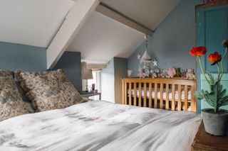 A double bed in a loft conversion with dark blue walls, a rose plant at the end of the bed, and a cot to the rear wall