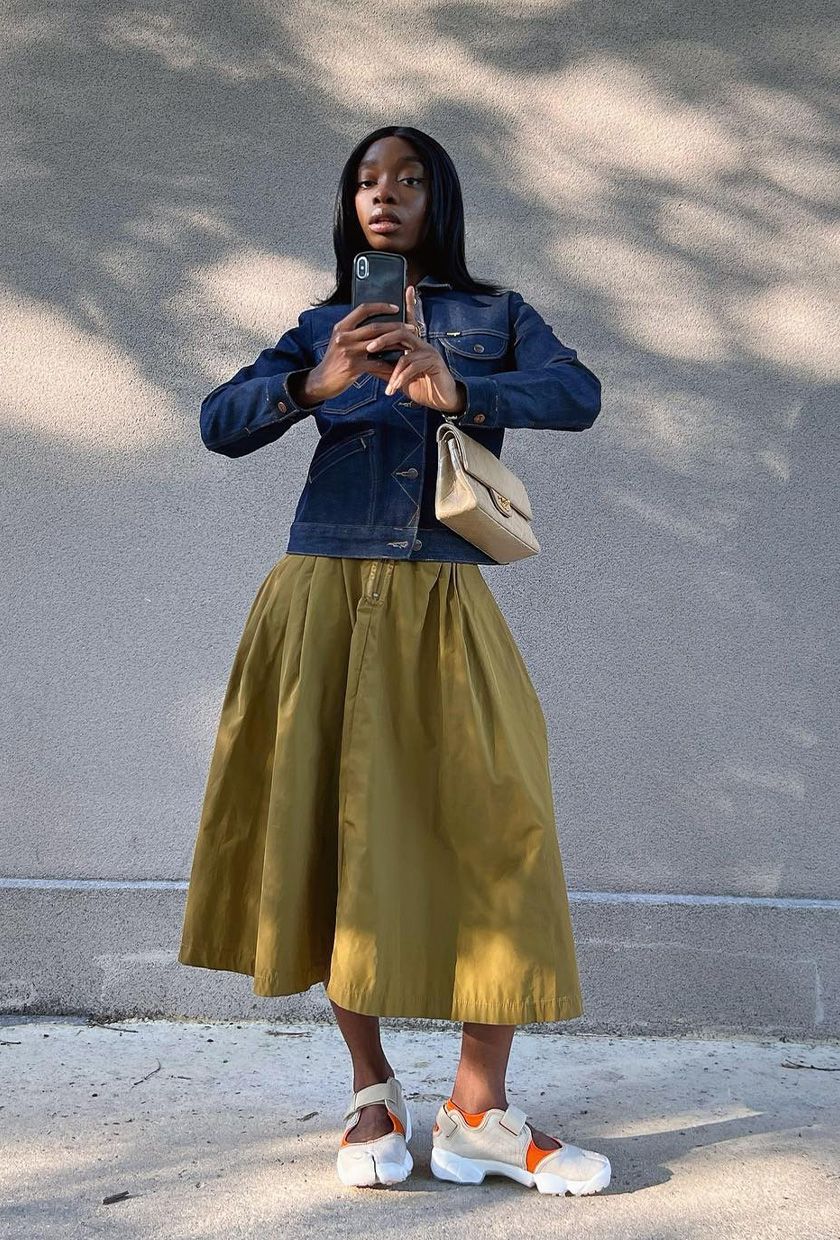 The best ballet sneakers are shown in a photo of a woman standing outside wearing a denim jacket with a green midi skirt and white mary jane sneakers