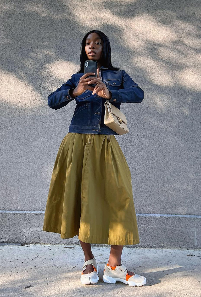 The best ballet sneakers are shown in a photo of a woman standing outside wearing a denim jacket with a green midi skirt and white mary jane sneakers