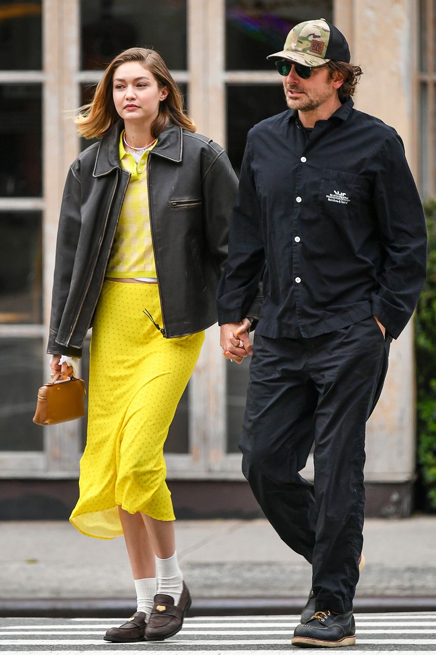 Gigi Hadid wearing a leather jacket and yellow polo and slip skirt with white socks and loafers, walking with Bradley Cooper