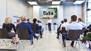 A woman leads a presentation in a full conference room in front of a Sharp display. 