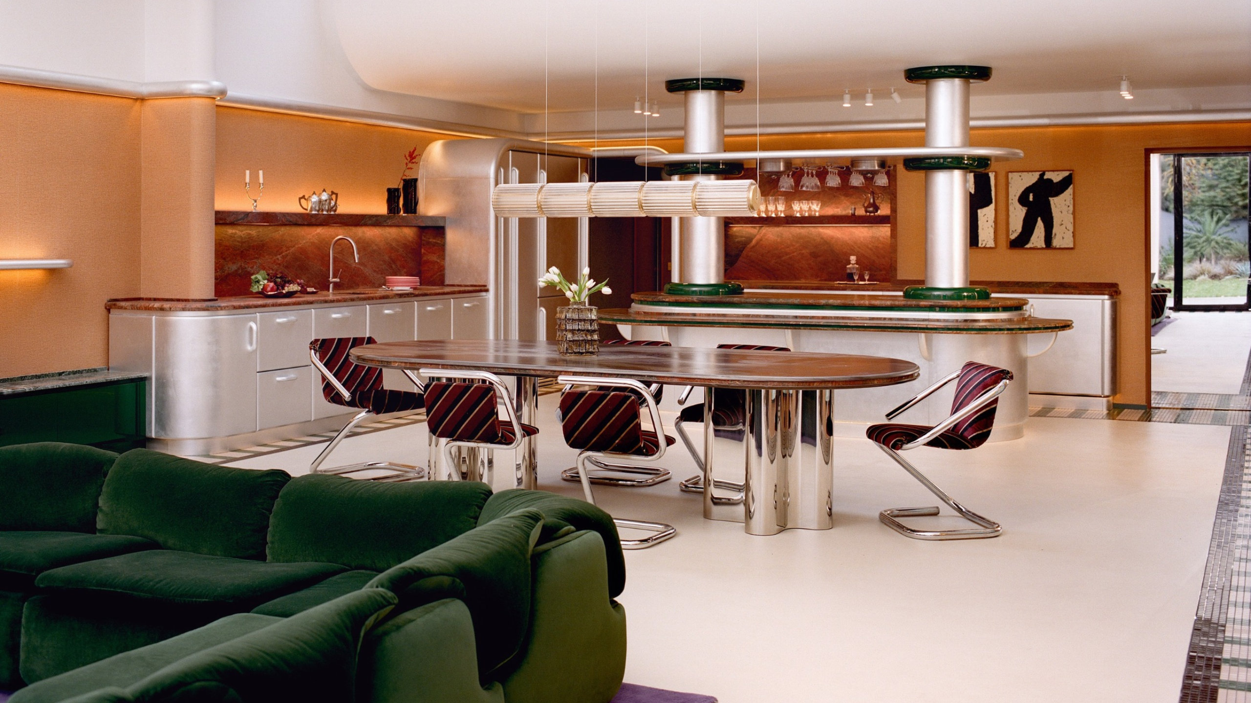 An open living, dining, and kitchen space in a Space Age-inspired home. There is orange wallpaper, a white ceiling, mosaic tiles floors, a dining table with purple chairs and a green sofa