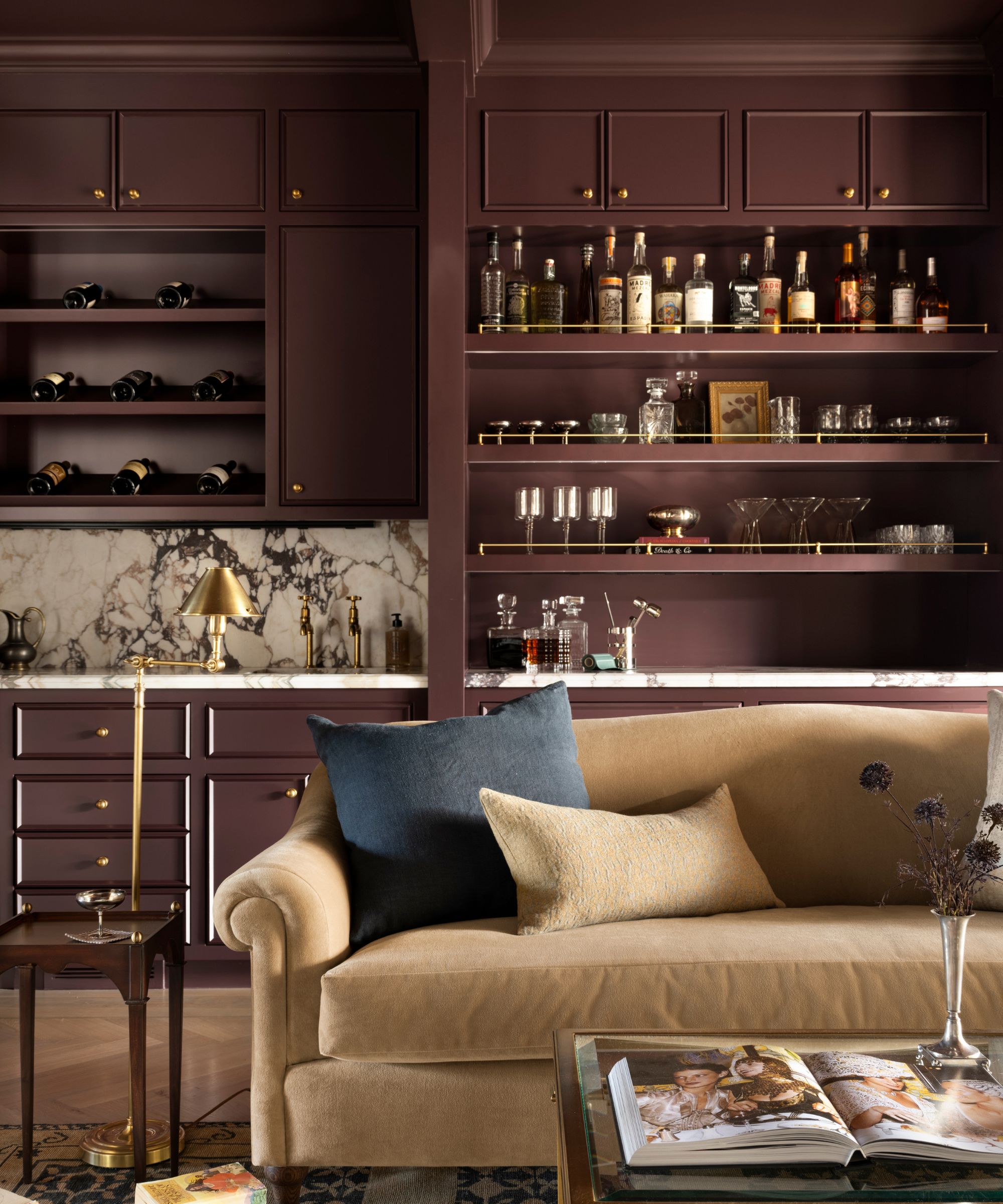 Living room with built-in home bar painted burgundy, shelves of glassware and alcohol, a marble countertop and backsplash, a velvet beige sofa, and a glass coffee table with an open book on top.