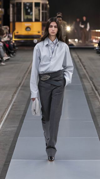 Tod's model wearing two button-down shirts layered on top of each other with a sideways belt and gray trousers.