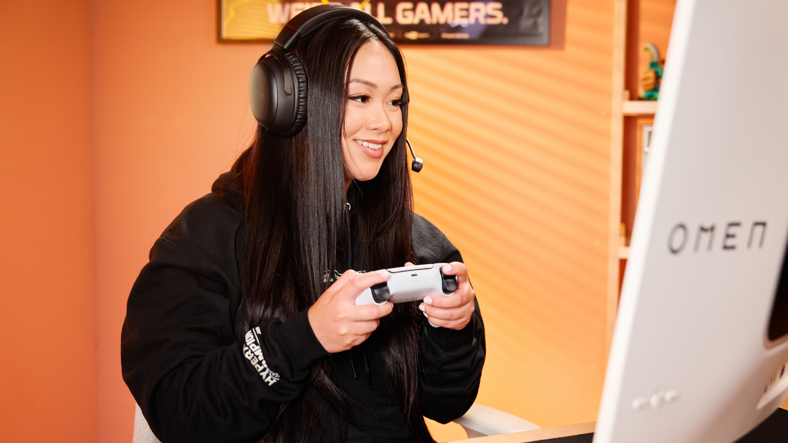 A woman with long dark hair and a black hoodie holds a controller while wearing a HyperX headset and gaming on an HP Omen monitor.