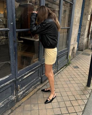 French influencer Anne-Laure Mais peeks into a window of a Paris shop while wearing a black leather jacket yellow miniskirt, and strappy black slingback flats