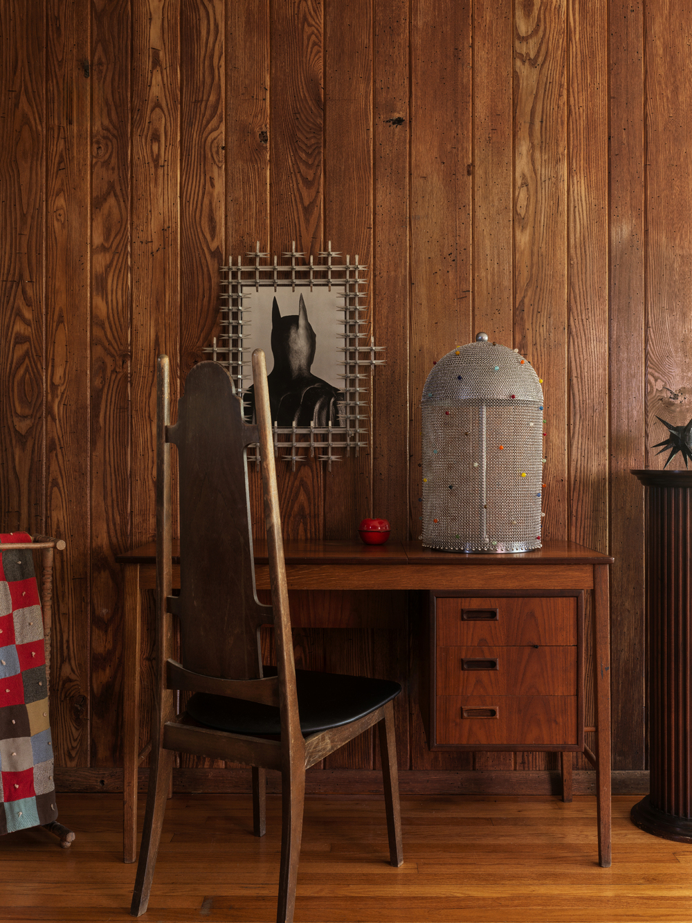 timber desk next to a timber panelled wall, with timber flooring, a chainmail lamp on the desk, a tall timber chair, and a framed artwork of batman in a spiky silver frame