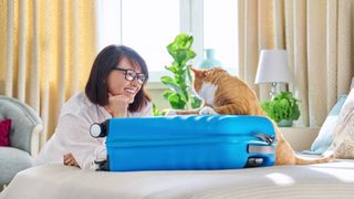 a woman smiles at her orange cat who has their front paws on a blue suitcase