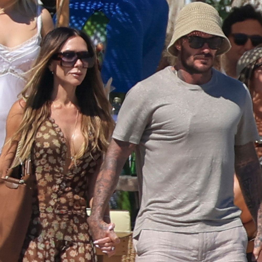Victoria and David Beckham walk along a beach holding hands. Victoria wears a brown sheer dress with Hermes Oran sandals whilst David wears a grey t-shirt with grey linen shorts.